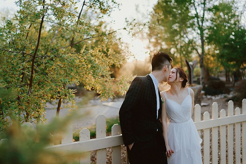 Wedding couple shares intimate moment by white picket fence during golden hour.