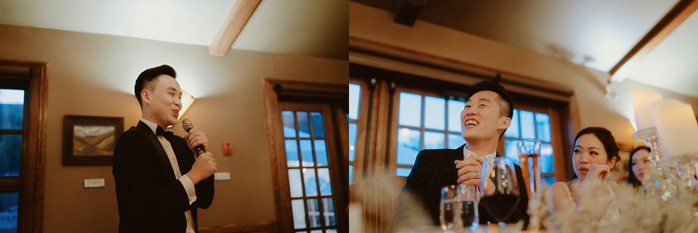 Wedding guests laugh during reception speeches in a rustic venue with wooden beams.