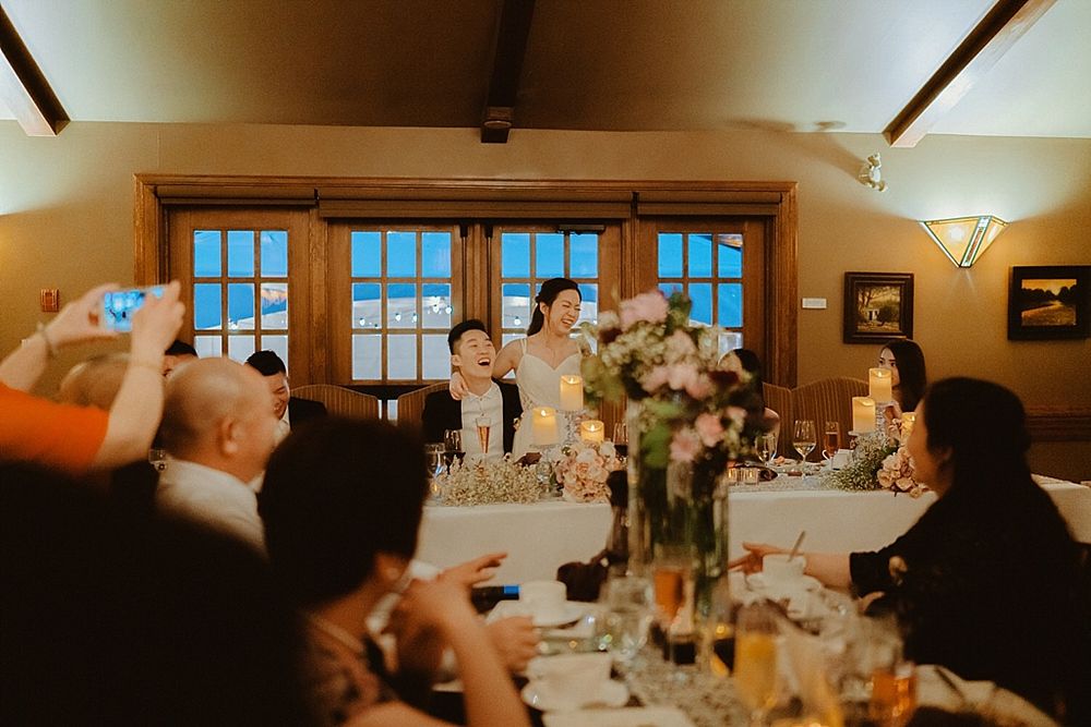 Guests enjoy dinner at an elegant wedding reception with floral centerpieces and large windows.