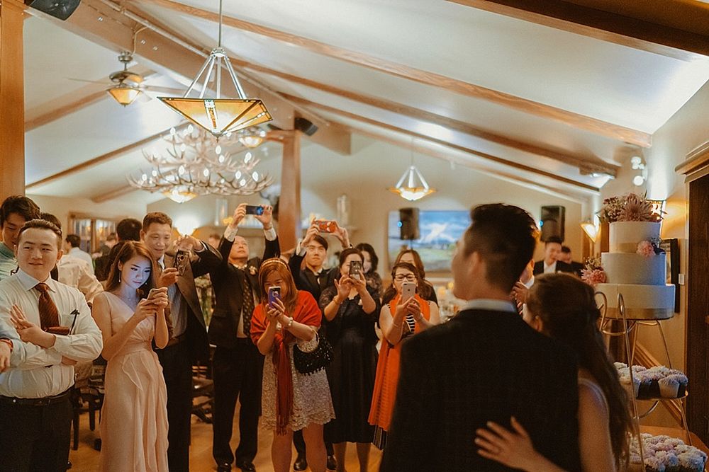 Wedding reception guests enjoying festivities under elegant chandeliers and wooden beams.