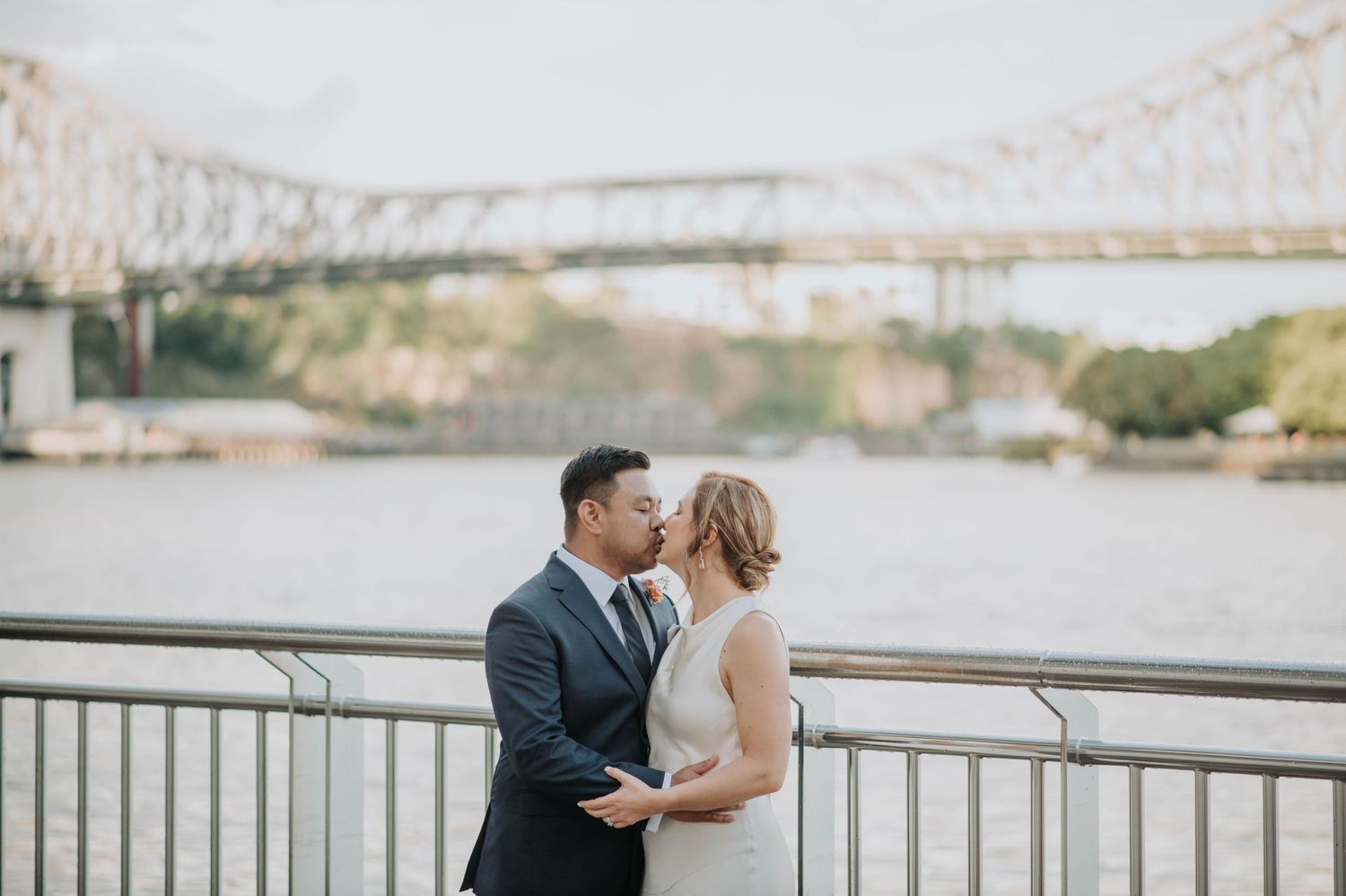 Couple embraces on riverside terrace with iconic bridge in background during wedding photoshoot.