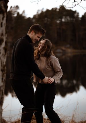 Laughing couple in dark winter coats share a joyful moment by a calm lake surrounded by trees.