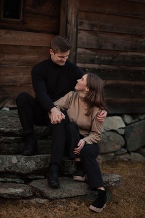 Couple in cozy sweaters sharing an intimate moment while sitting on rustic stone steps by a wooden cabin.