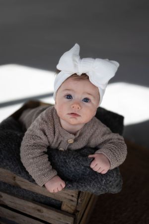 Baby wearing white bow headband and gray sweater lying on dark cushion in wooden crate.