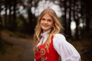 Portrait in traditional Norwegian folk costume with red vest against forest background.