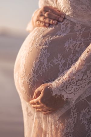 Close-up of hands cradling a pregnant belly covered in delicate white lace against a sunset backdrop.