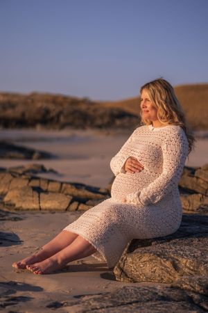 A serene beach maternity portrait features a person in white lace dress seated on coastal rocks.