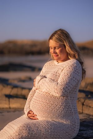 A person in a white knit dress sits peacefully on rocks at the beach during golden hour.