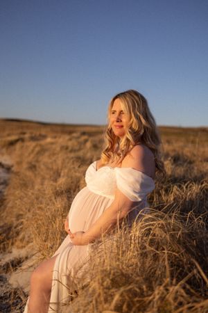 Pregnant woman in white dress poses in golden field during sunset photoshoot.