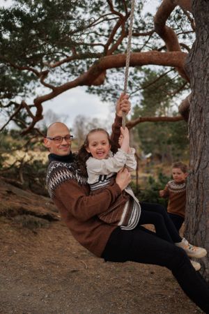 A playful scene shows two people swinging from a tree branch while wearing knit sweaters in an outdoor setting.
