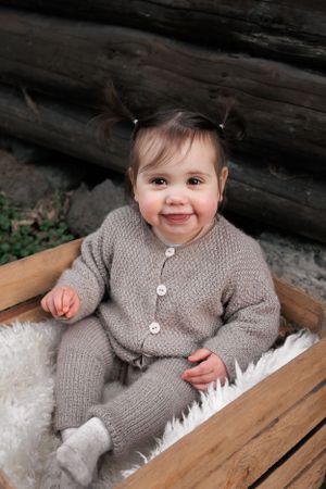 A baby wearing a cozy grey knit cardigan and pants sits on a wooden bench with white fleece.