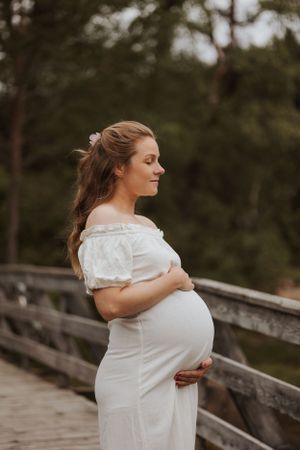 An elegant maternity portrait showing a woman in white dress posing in profile on a wooden bridge.