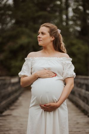 A maternity photo of an expectant mother in a white off-shoulder dress standing on a wooden bridge.