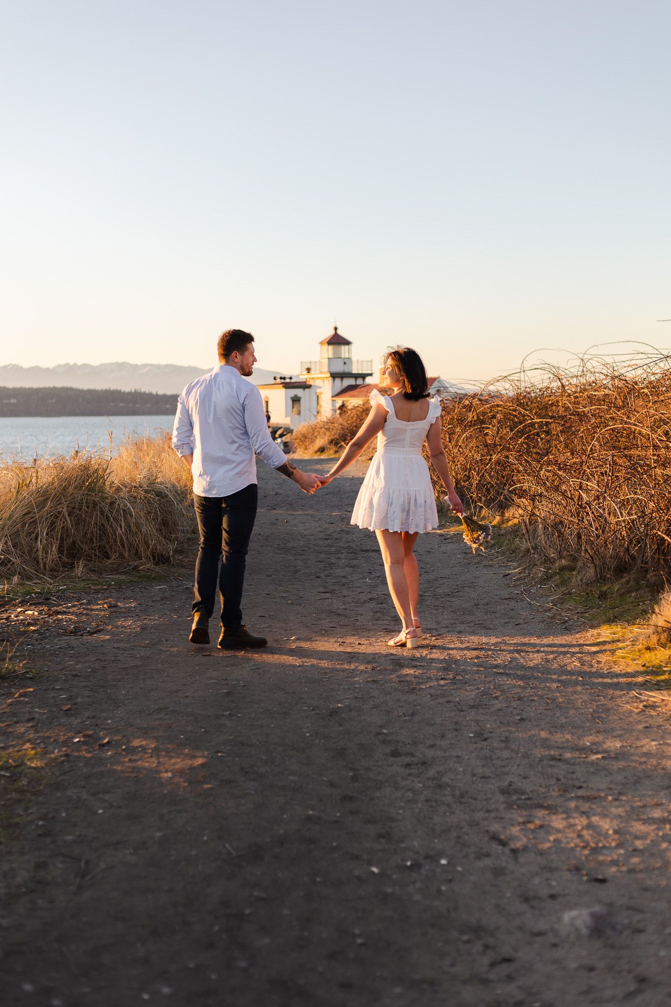 A romantic couple walks along a coastal path with a lighthouse in the distance at sunset.