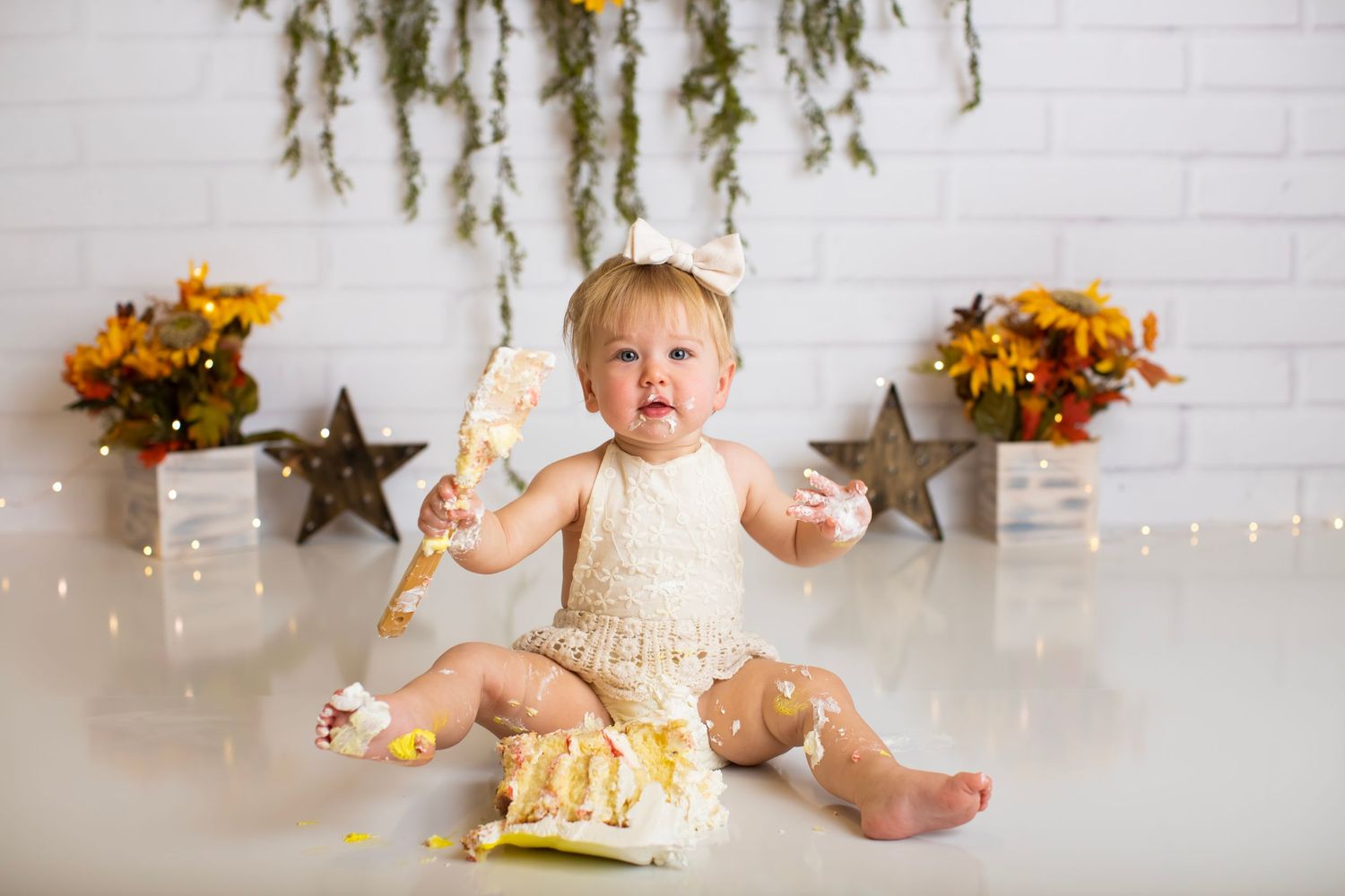 Baby in cream dress surrounded by rustic decor and sunflowers for cake smash session.