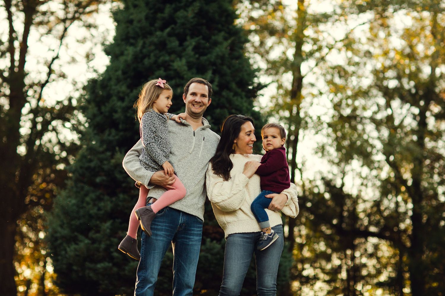 Family of four poses together outdoors against evergreen trees at sunset.