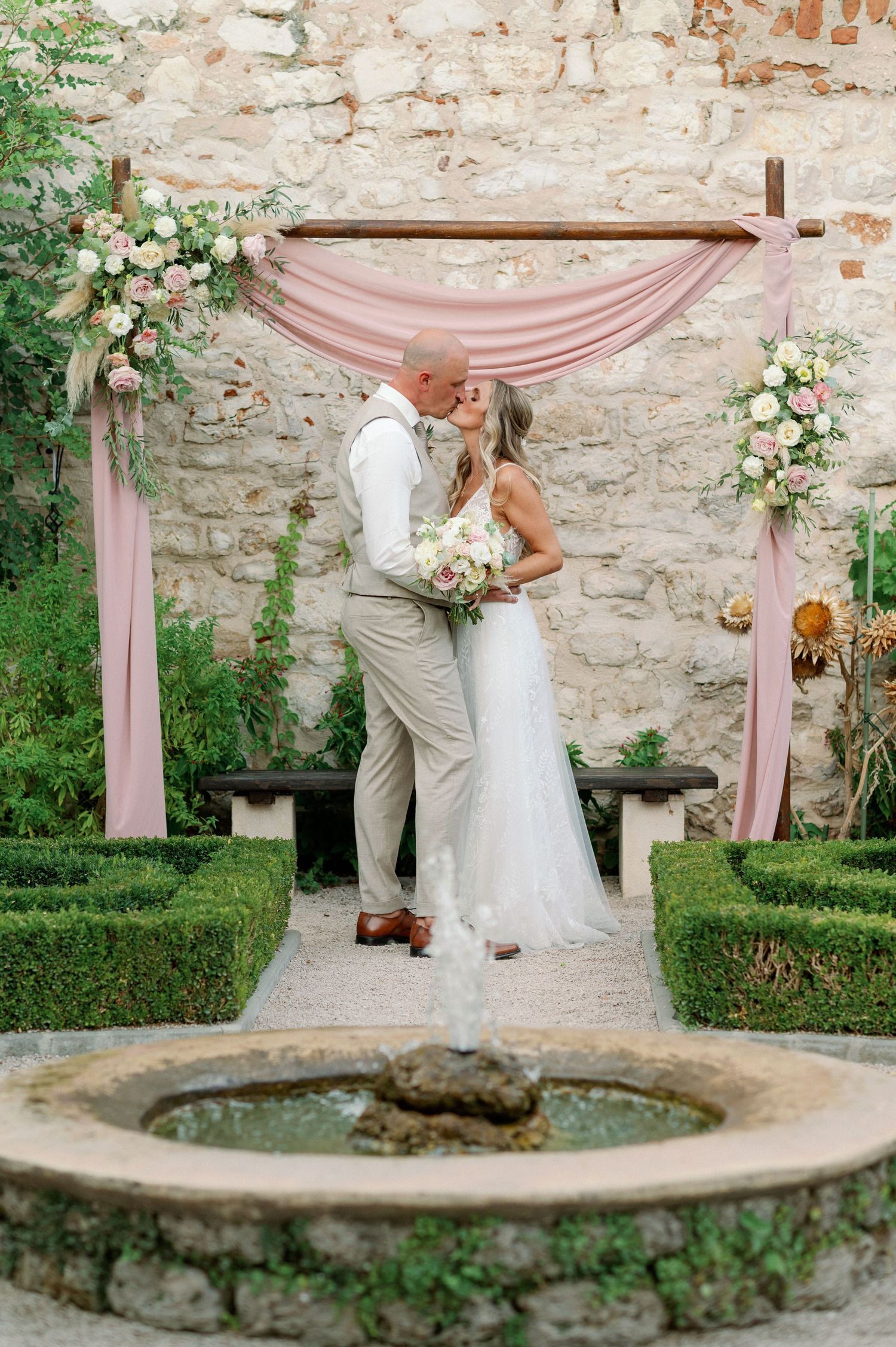 Couple embraces under pink draped arch with flowers in a garden courtyard with stone walls and fountain.