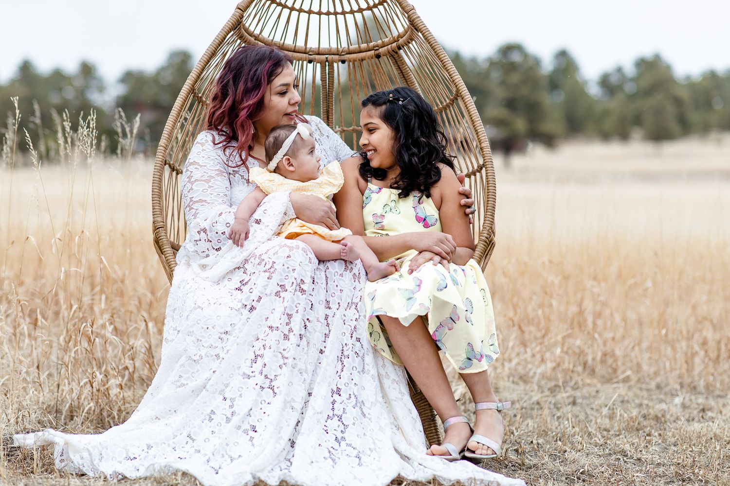 Mom holds her baby girl and sits next to her older daughter in an egg chair in Black Forest, Colorado.