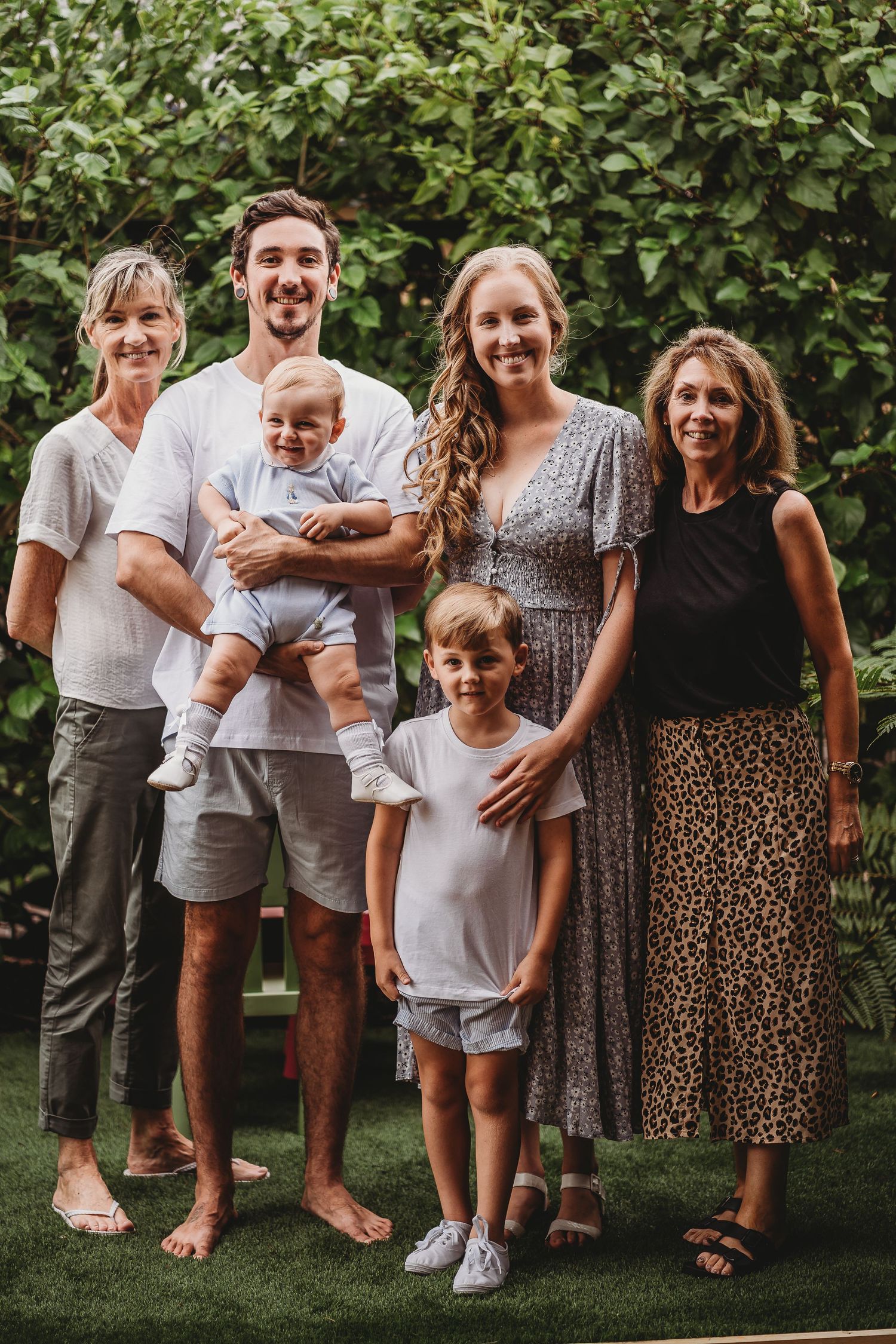 Group portrait of extended family members standing together outdoors on grass with greenery background.