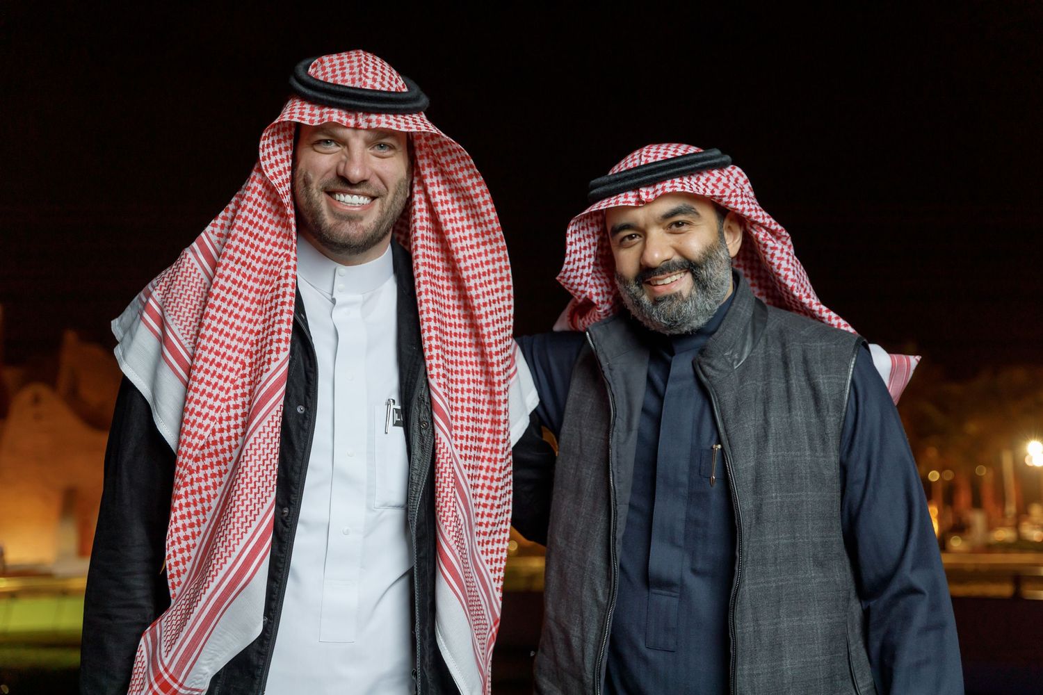 Two men in traditional Middle Eastern attire pose together at night with buildings in background.