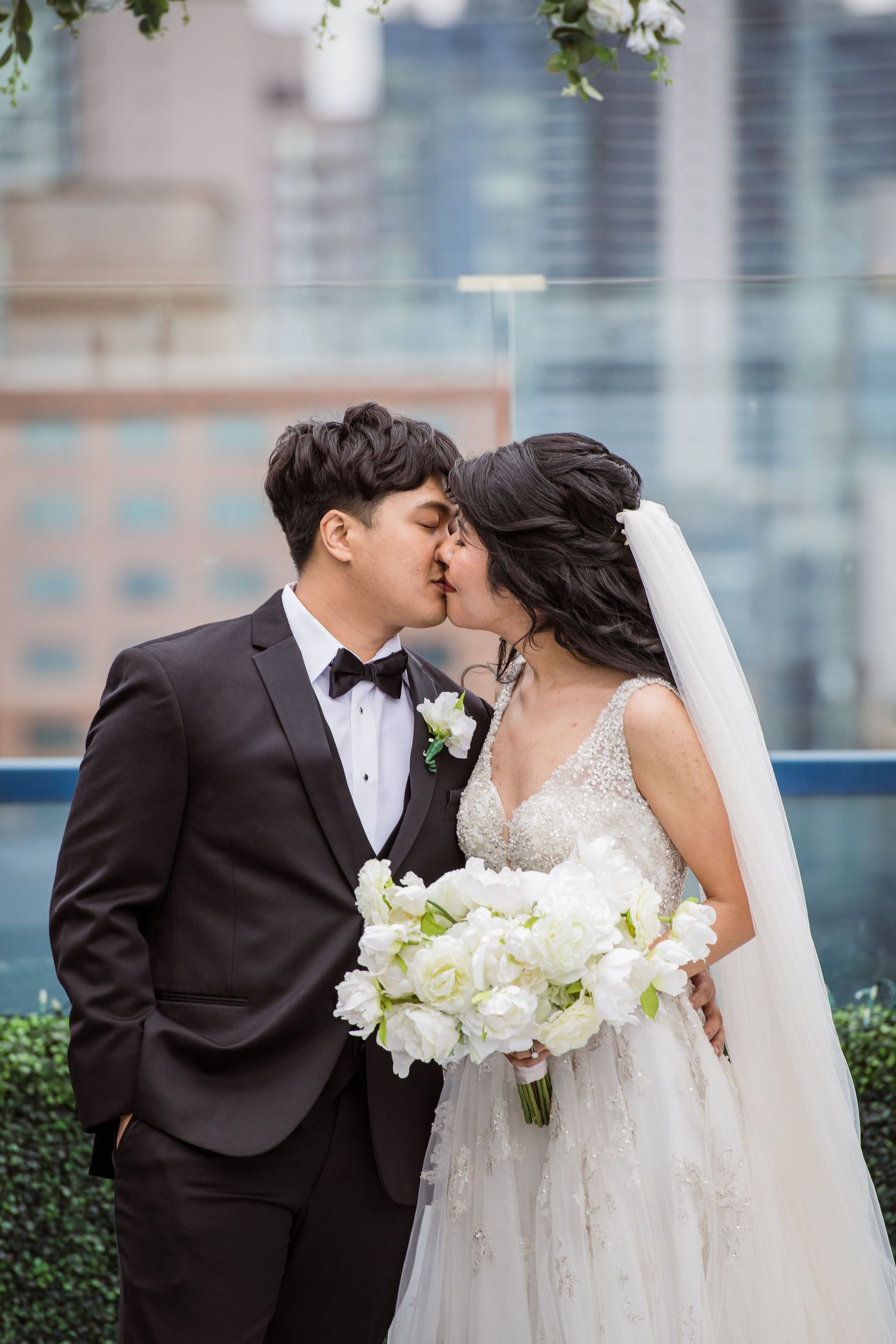 Asian bride and groom sharing a kiss on a rooftop with a city skyline backdrop on their wedding day.