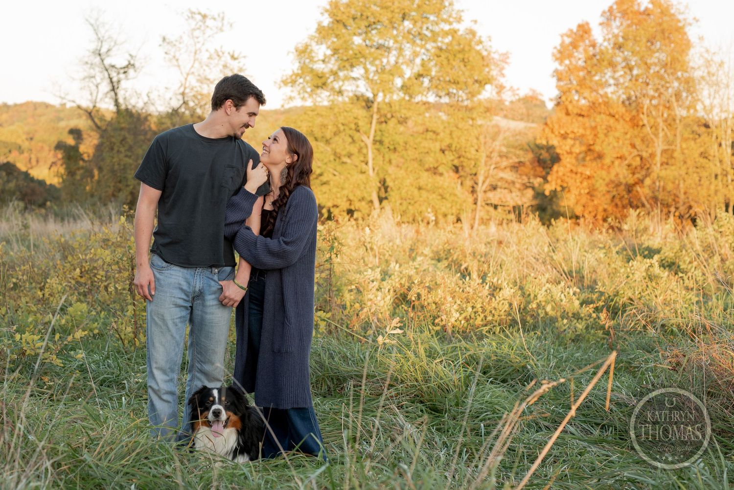 A couple stands together with their Australian Shepherd dog in a golden autumn field.