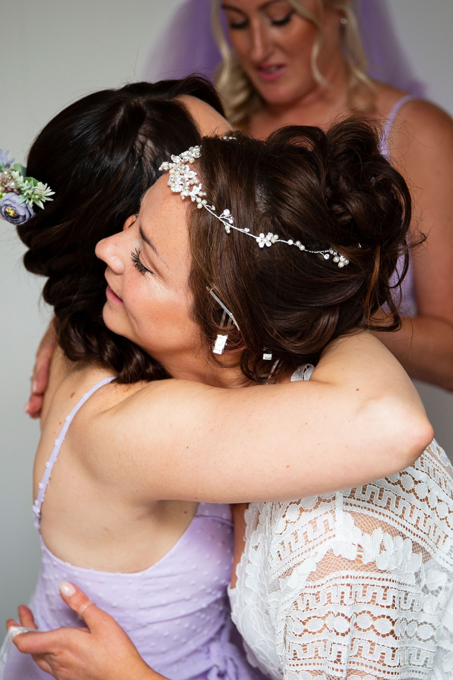 Bride in a white lace dress embraces bridesmaids wearing lavender dresses during an emotional wedding moment.