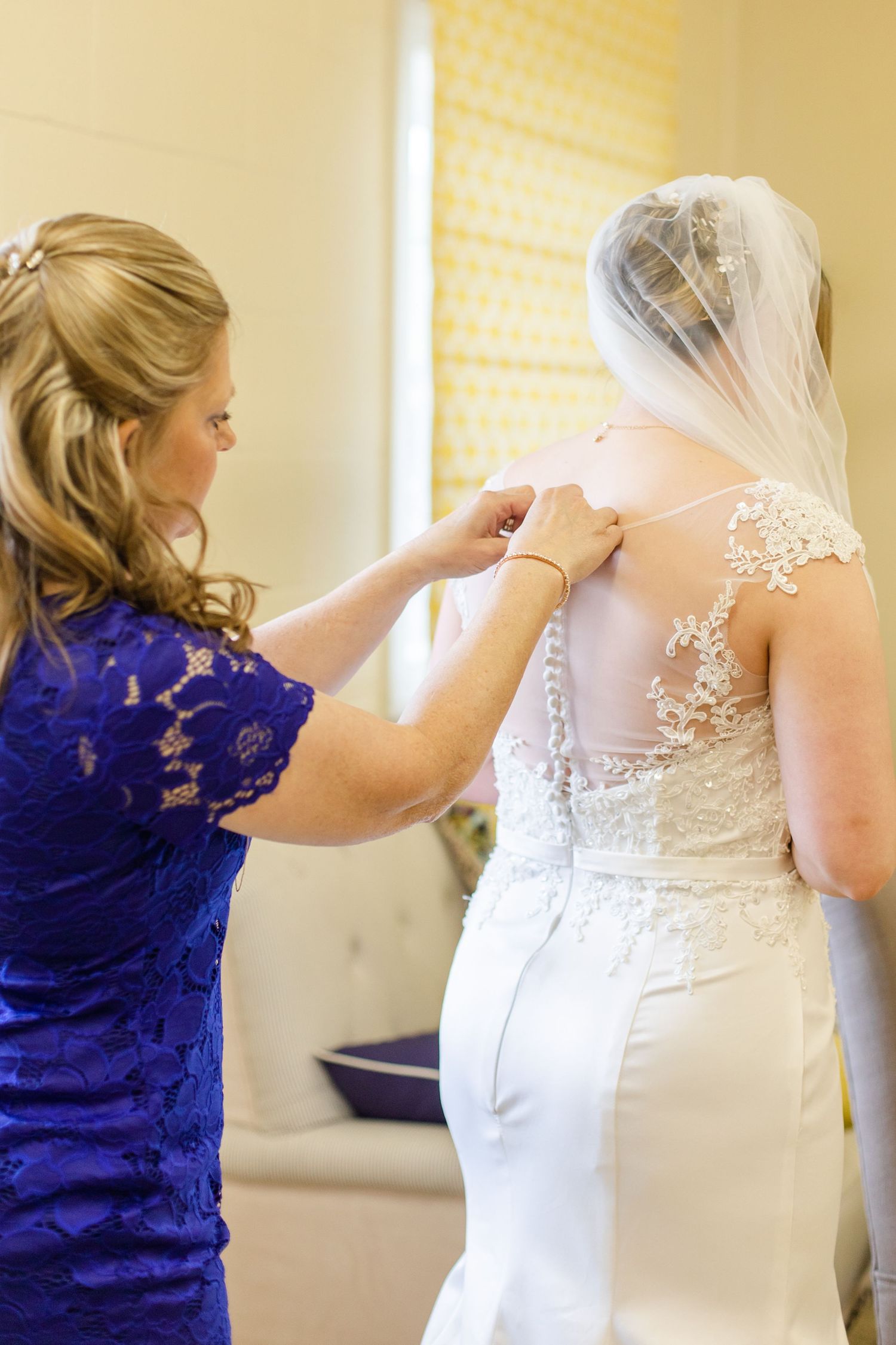 A bridesmaid in royal blue helps adjust the back of a white lace wedding gown.