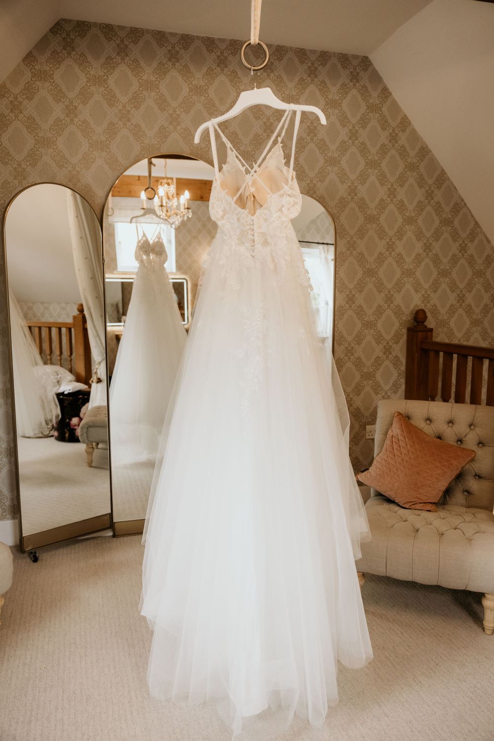 A delicate white lace wedding gown hangs in front of an ornate mirror and patterned wallpaper.