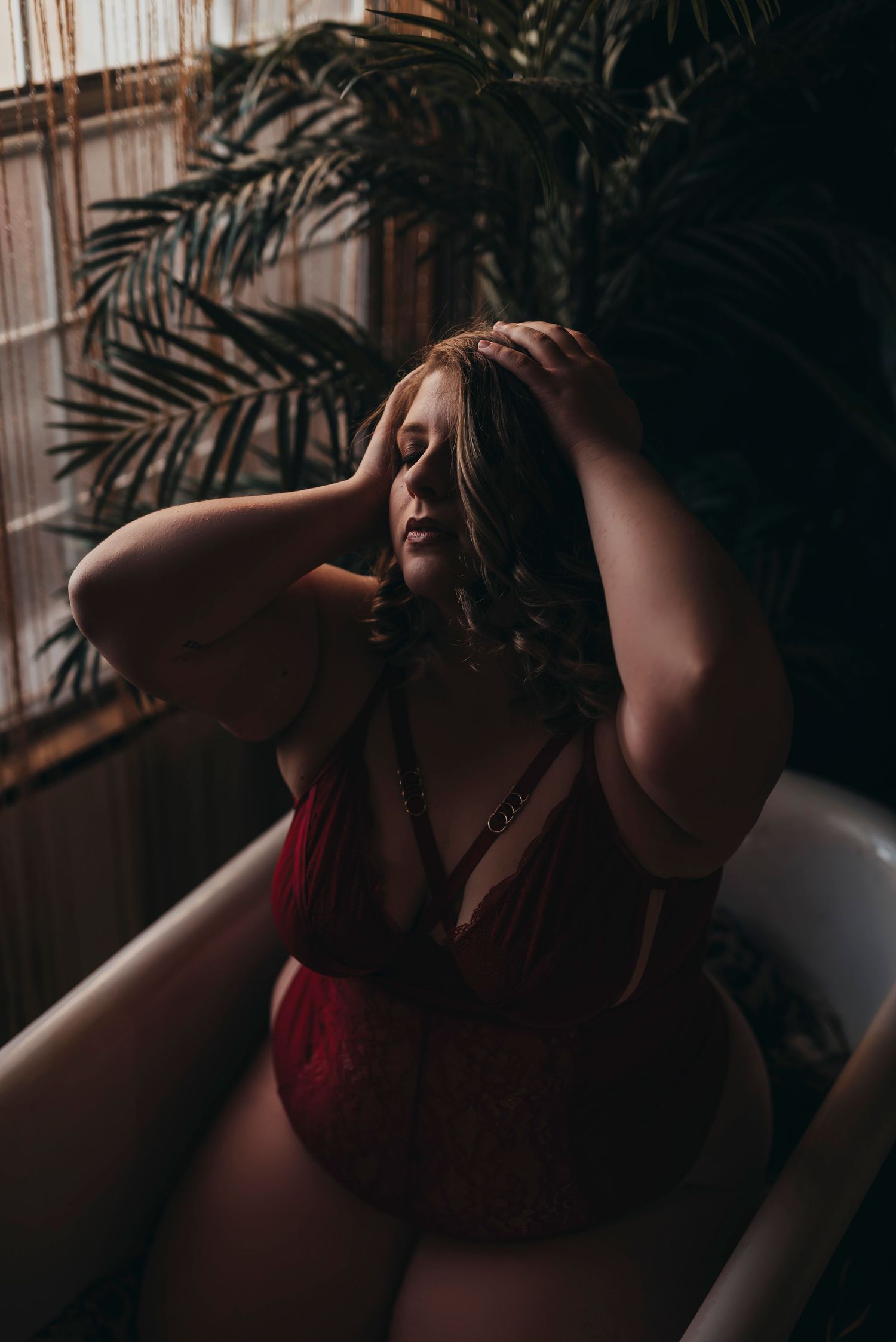 Moody boudoir photo in dark room with red lingerie and dramatic palm plant shadows by window.