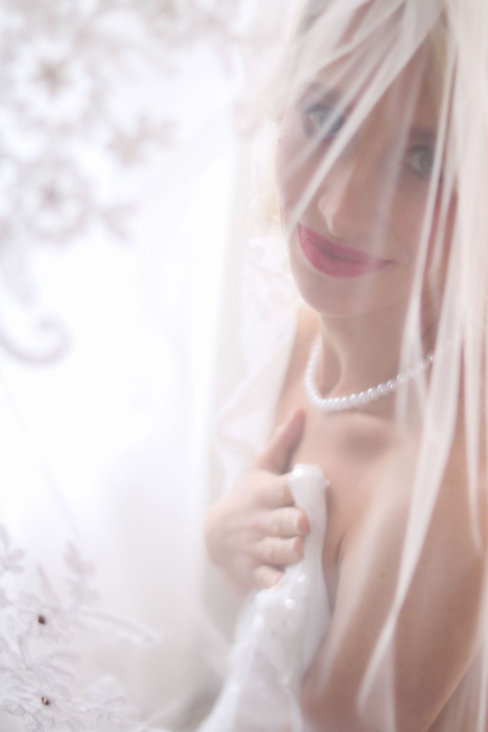 Soft ethereal bridal portrait through delicate white veil showing romantic makeup and pearls.