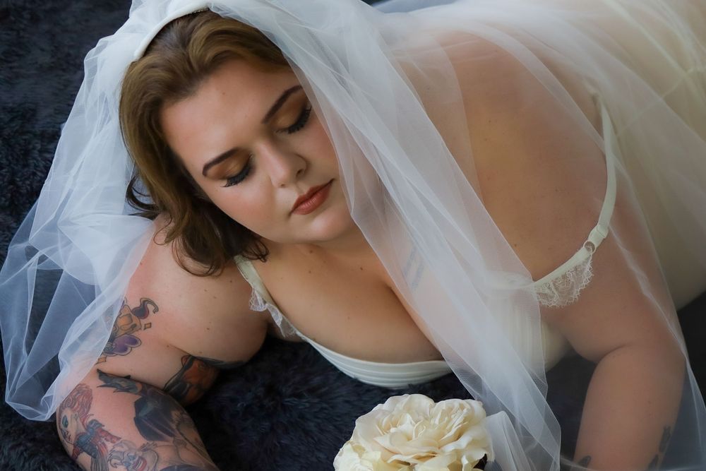A bride in lingerie and veil poses with white roses against a dark background.