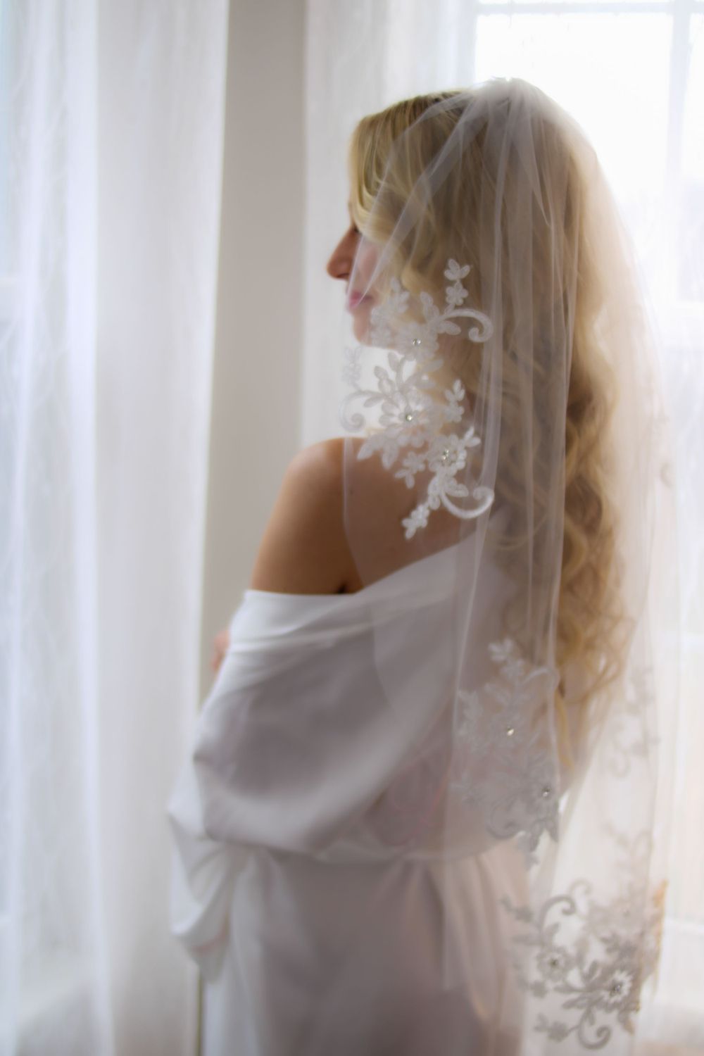Person in white robe with lace veil by window.