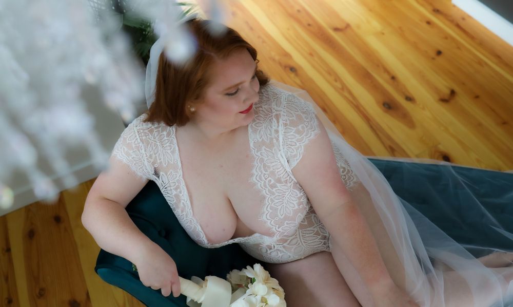 A bride in a white lace gown with a bouquet poses on a wooden floor.
