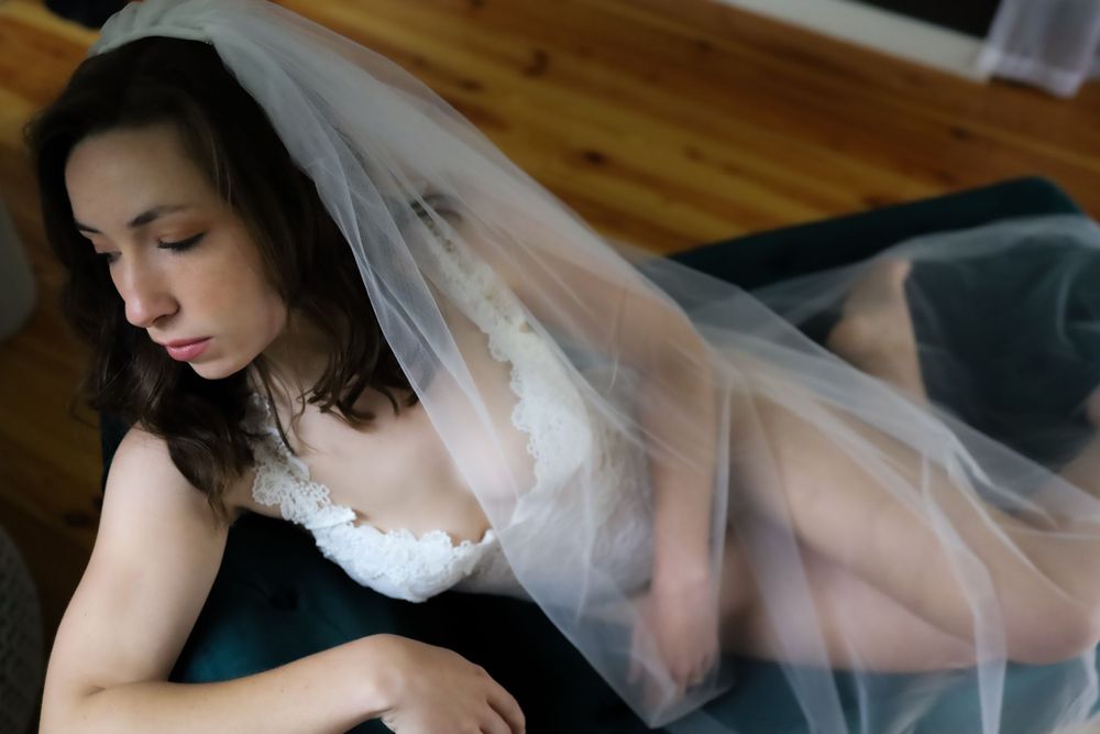 A bride in white lace and flowing veil reclines in an artistic boudoir pose.