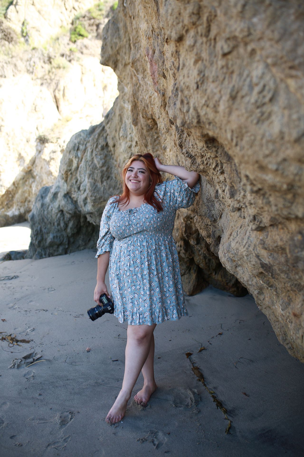 Person in floral dress poses barefoot against rocky beach cliffs.