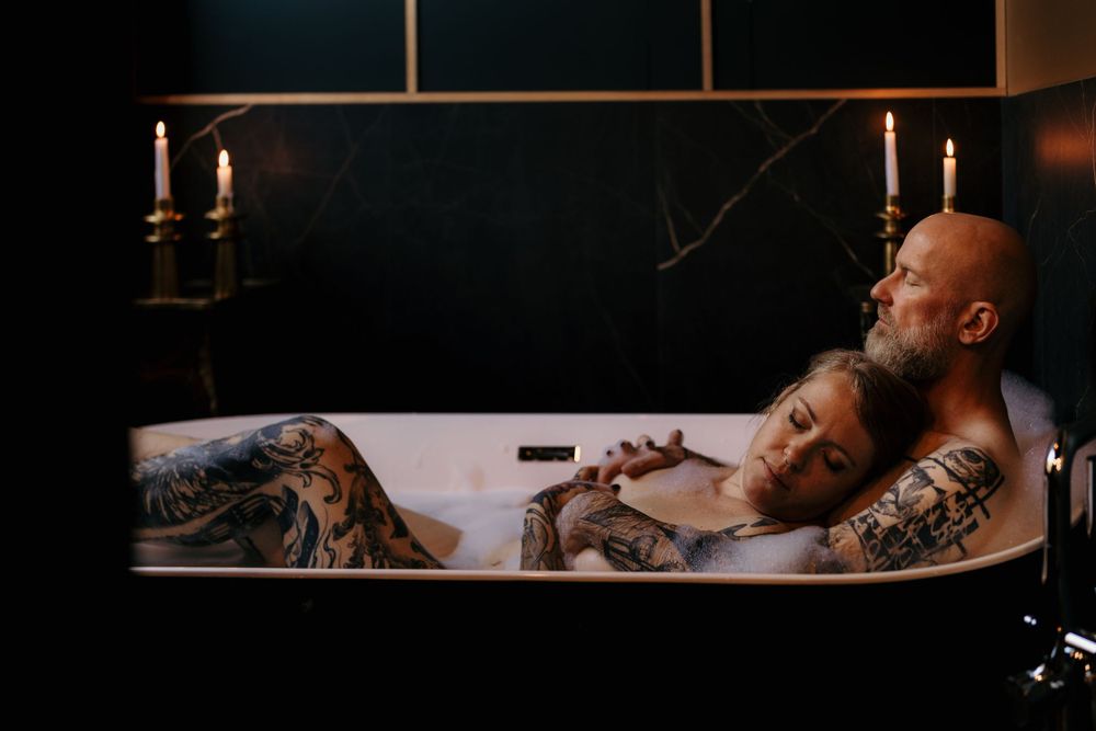 A tattooed couple shares an intimate moment in a bathtub lit by candlelight.