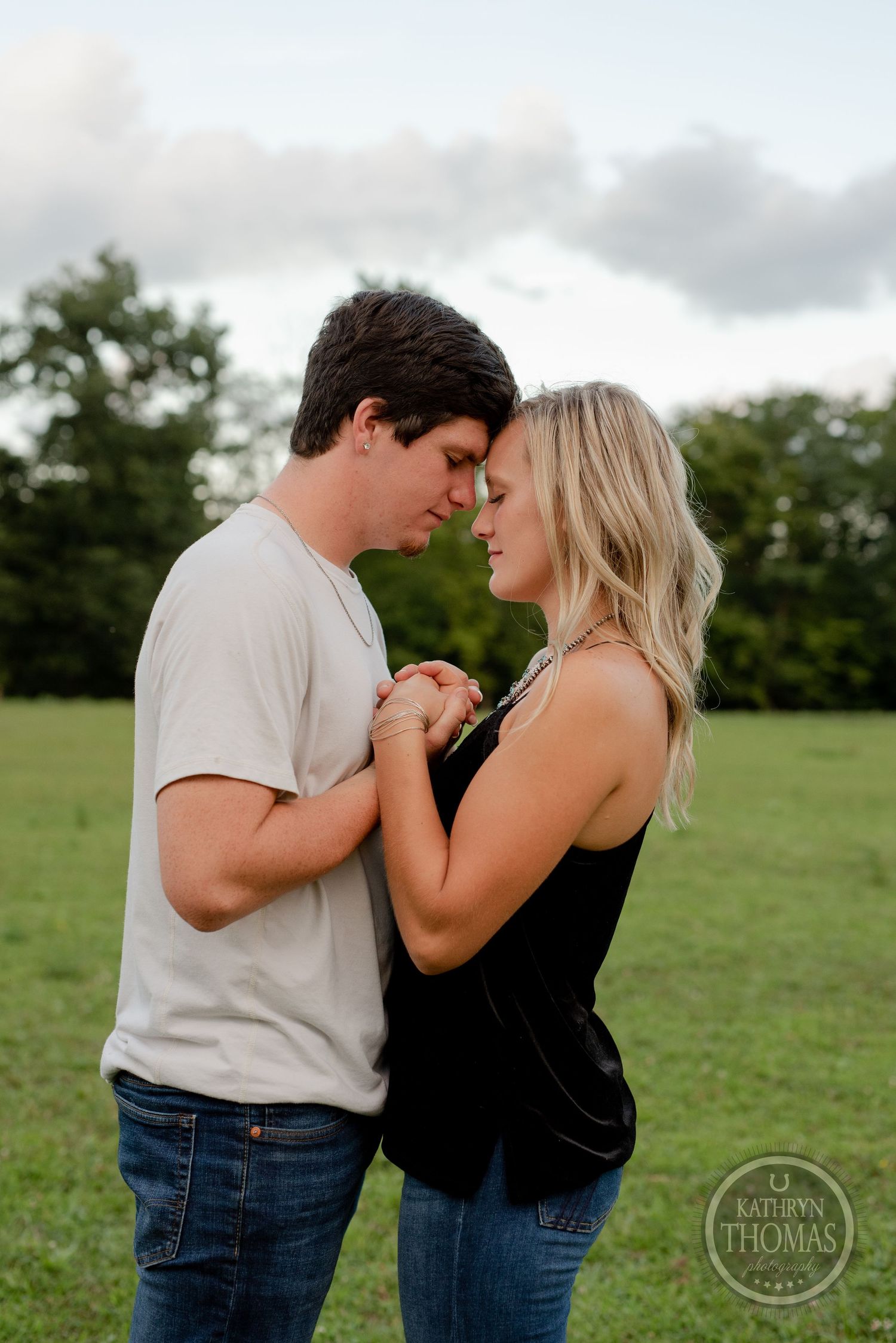 A romantic couple shares an intimate moment while holding hands in a green field at dusk.