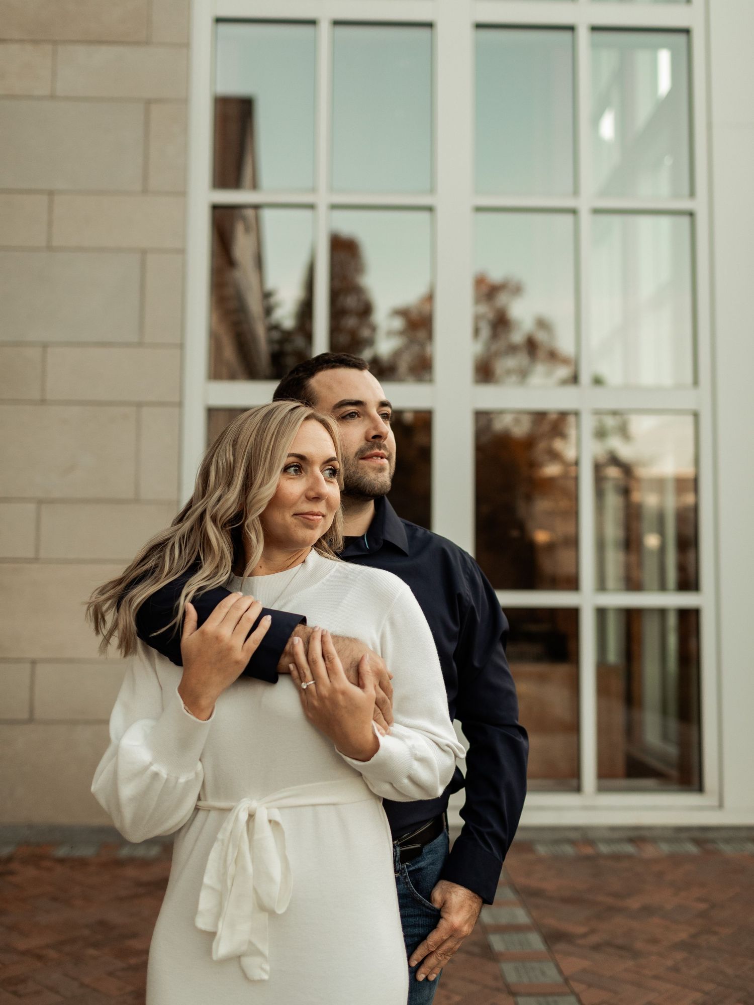 Couple posing together against white brick wall with large window panels.