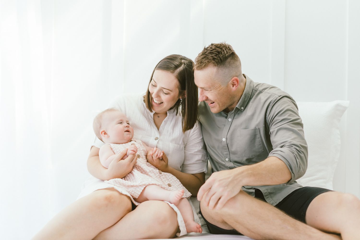 Parents cuddle and play with baby during intimate lifestyle photography session on white bedding.