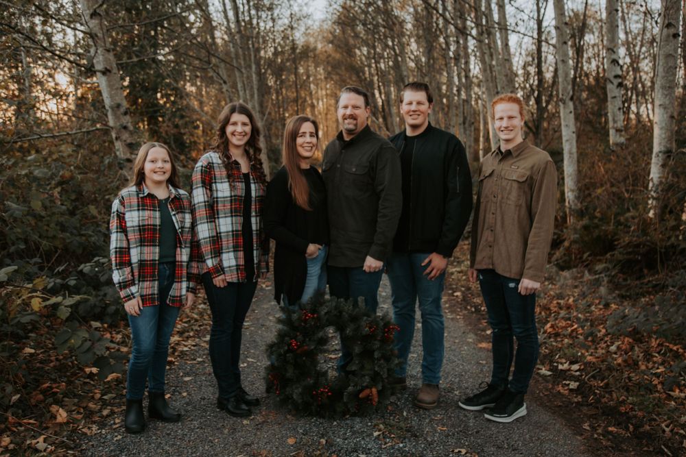 Family of six in plaid and neutral fall outfits poses on a forest path behind a Christmas wreath.