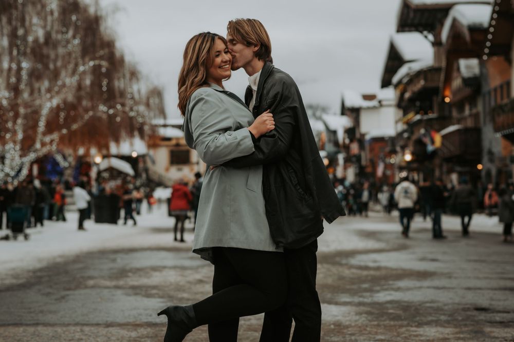 Couple in winter coats embrace and kiss on a snowy street in a charming alpine village with shops and strolling visitors.