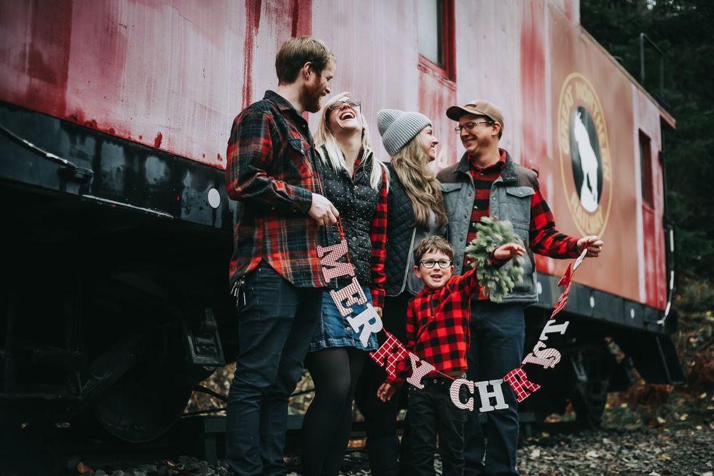 Family in plaid holds a Merry Christmas banner beside a vintage red train caboose in the woods.