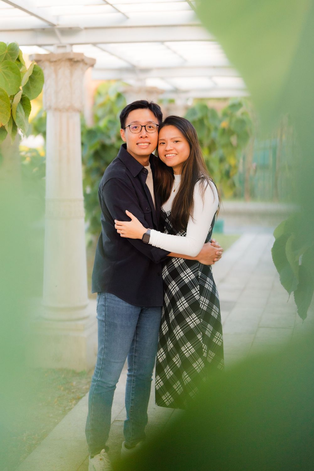A romantic couple sharing an embrace in front of a white column with greenery in the background.