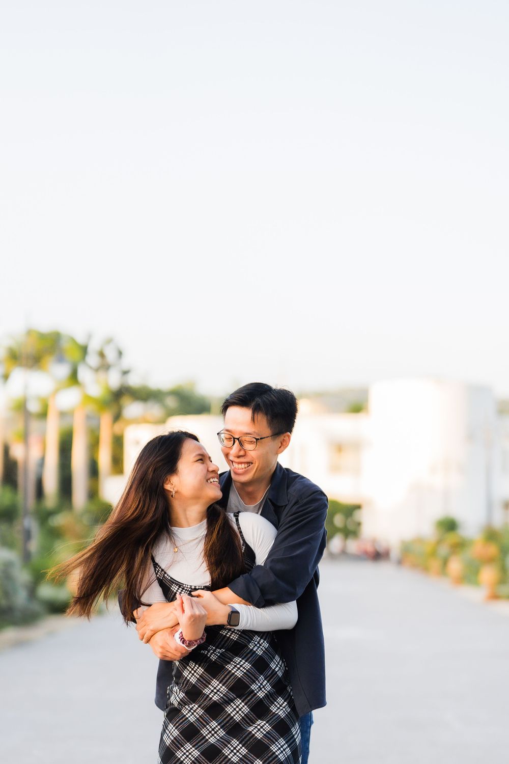 A happy couple embracing and laughing together on a sunny pathway with palm trees in the background.