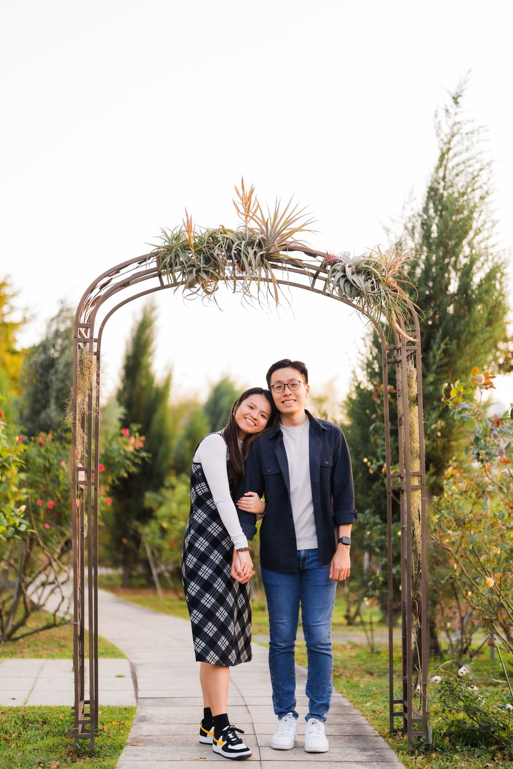 A couple stands together under a rustic garden arch with climbing plants.