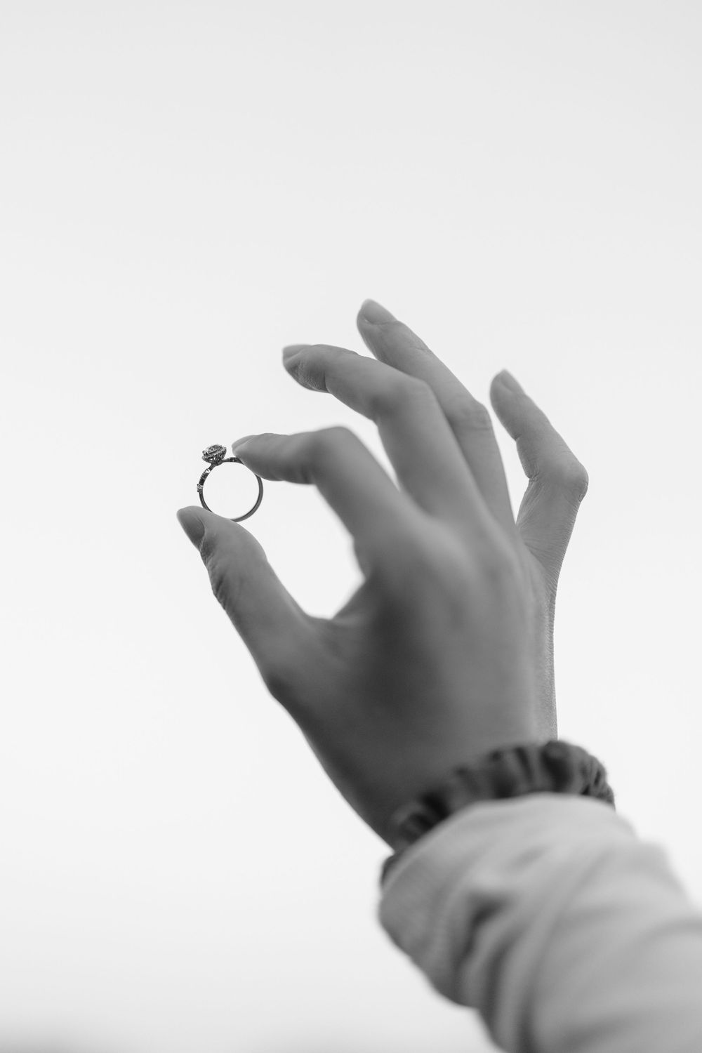Black and white close-up of hand holding engagement ring against light background.