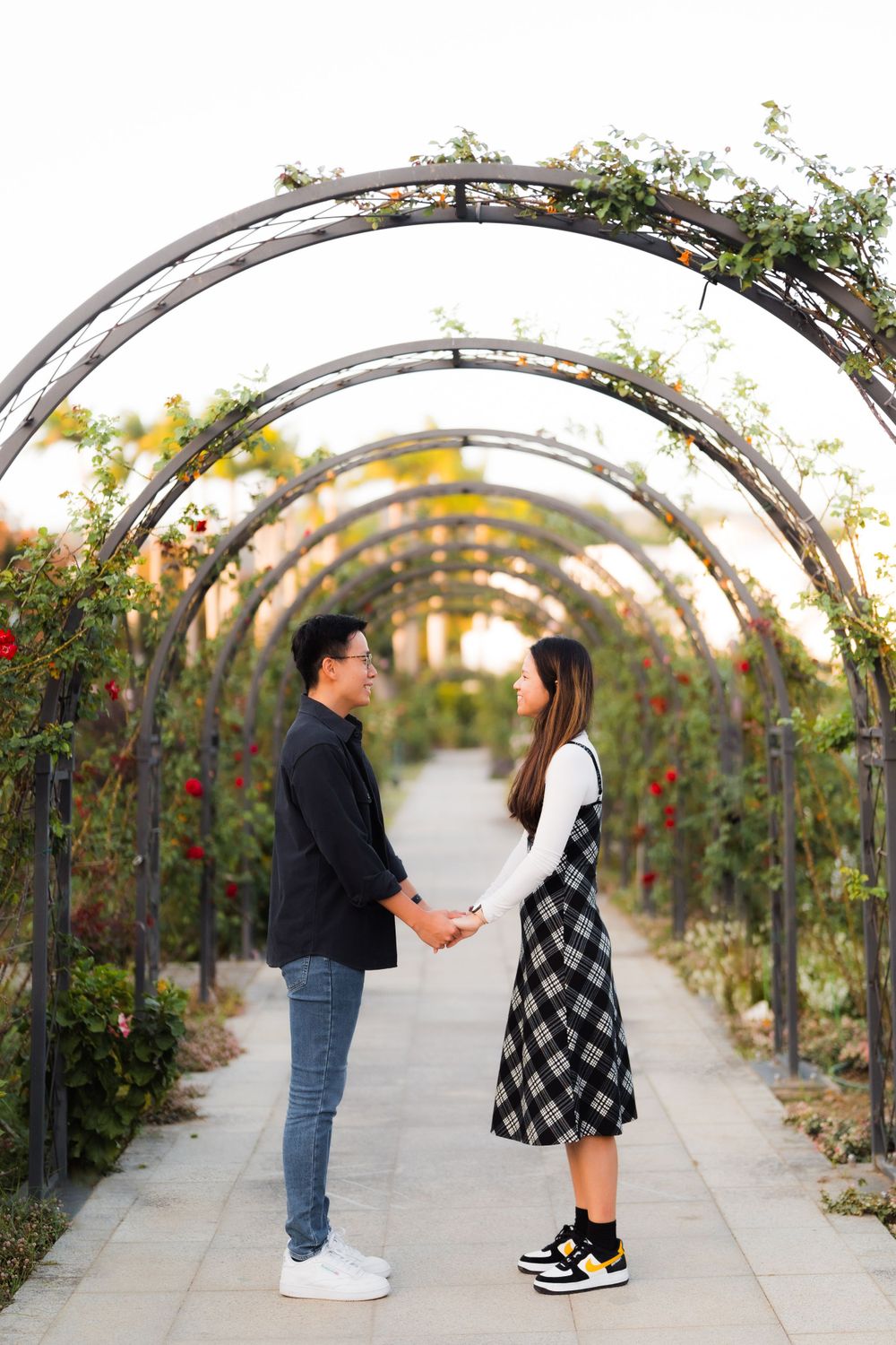 A couple stands face to face under an arched garden trellis walkway with climbing vines.