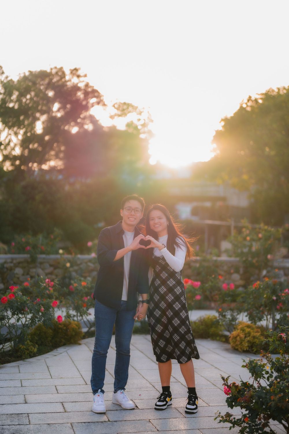 A couple making a heart shape with their hands against a sunset in a rose garden.