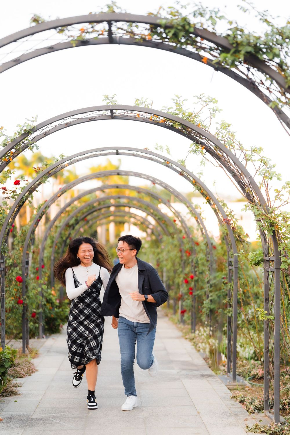 A couple running joyfully through an arched garden tunnel covered in climbing vines and flowers.